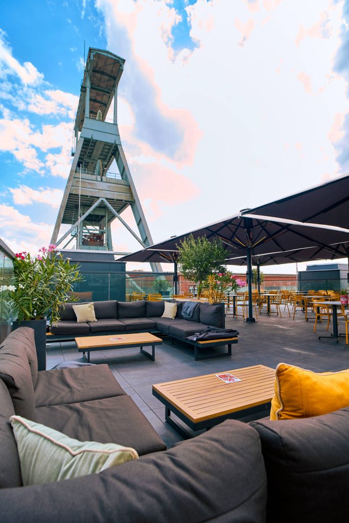 Außenterrasse mit Sofas, Tischen und Sonnenschirmen vor einem hohen Industriekran unter blauem Himmel